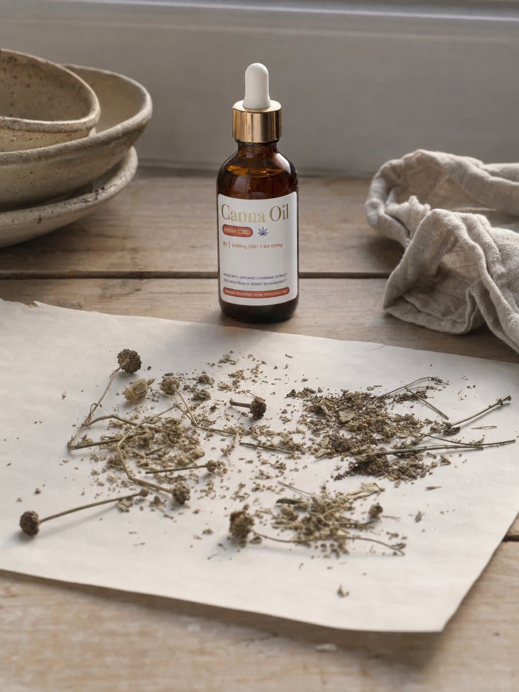 Dried whole botanical material, including stems, seed heads and crumbled leaf, spread across uncoated paper with the bottle behind it.