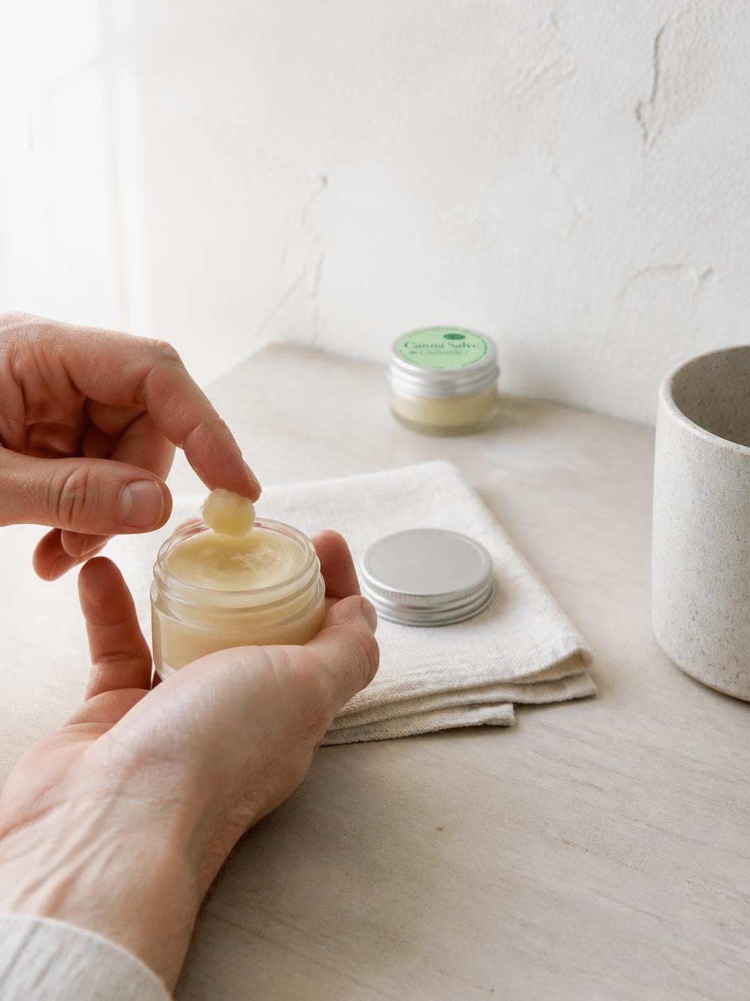 Two hands over a pale timber table in bright morning light, one holding an open glass jar of ivory balm and the other lifting a small soft scoop of it on a forefinger, the silver lid lying face-down on a folded cream linen cloth, a second closed jar with a pale green lid label standing out of focus behind, and a speckled stoneware cup at the right.