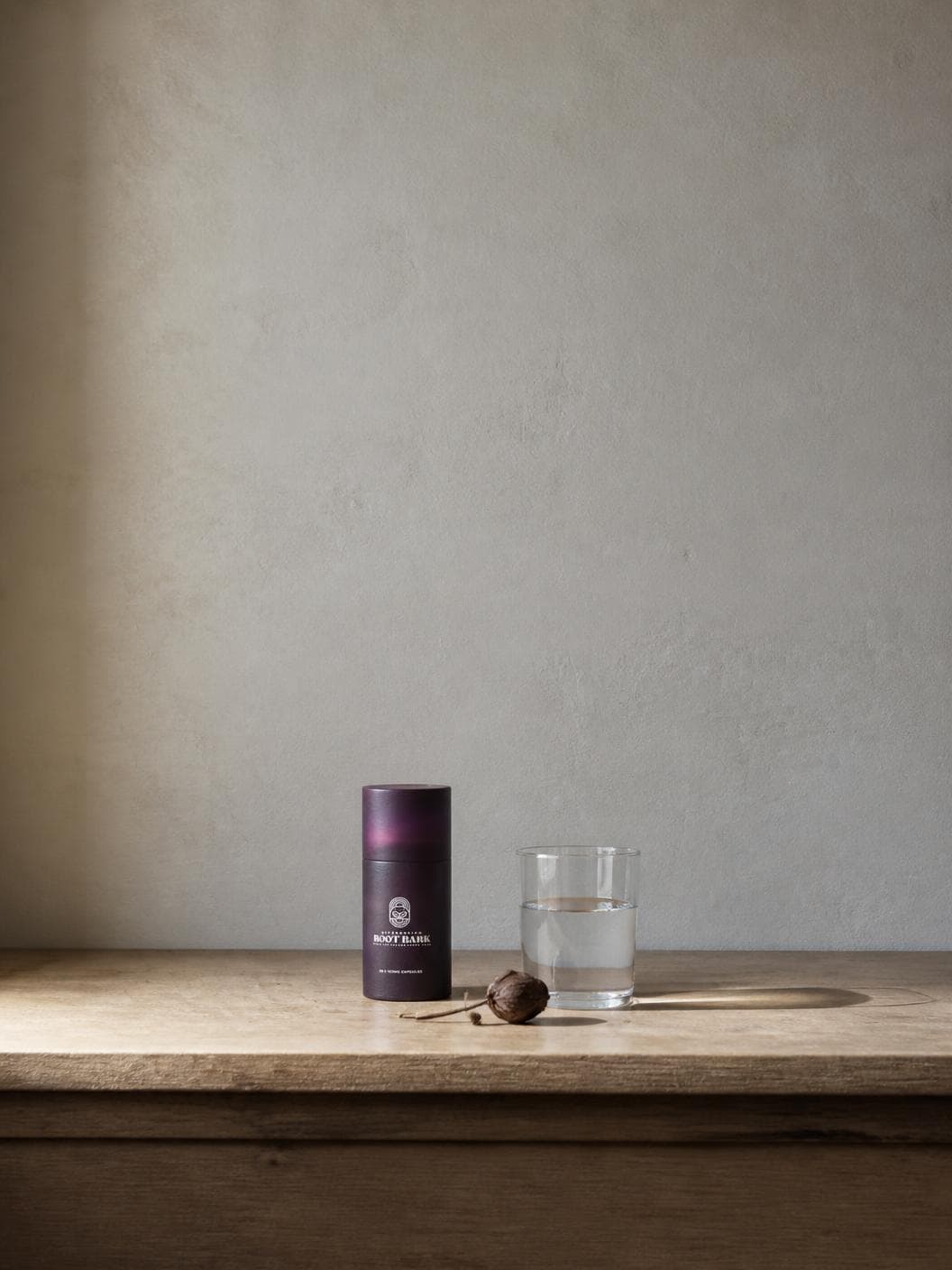 A bare timber sill below a wide, empty plastered wall in flat, still daylight: the closed aubergine tube standing small and low in the frame beside a plain glass of water and a single dried seed pod, and nothing else in the room.