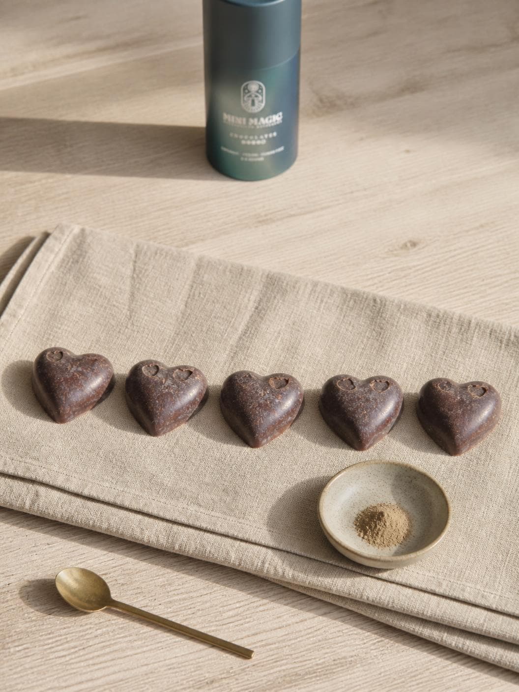 Five dark chocolate hearts in a row on folded oat linen over pale timber, each dusty with cocoa bloom, a small shallow ceramic dish of finely milled pale brown powder below them, a long brass spoon on the wood, and the petrol blue tube standing out of focus behind.
