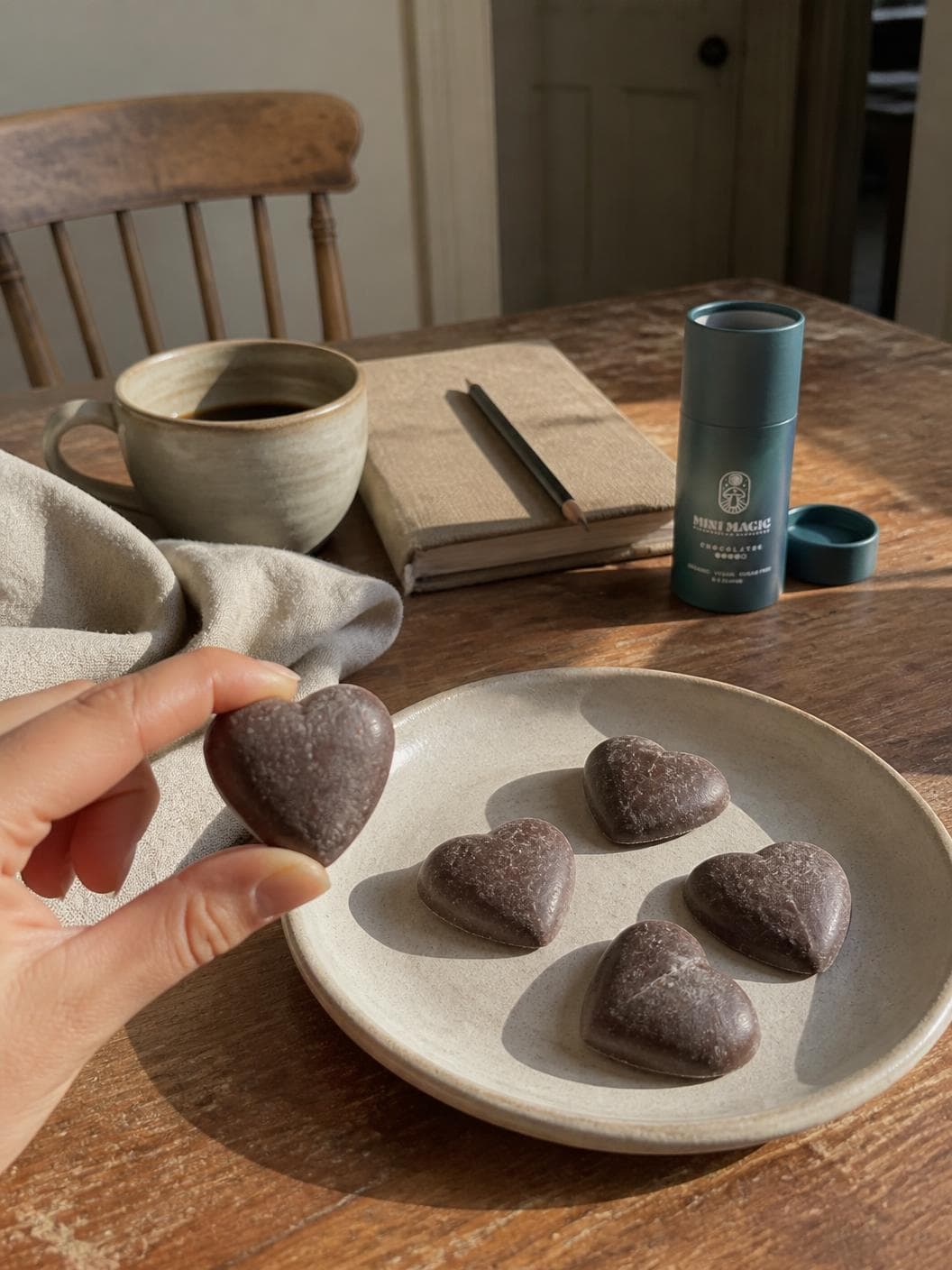 A hand at the left of a worn kitchen table holding a single chocolate heart between finger and thumb, a shallow stoneware plate of four more in front of it, a mug of black coffee and a clothbound notebook with a pencil across it behind, the petrol blue tube standing open beside its lifted lid, and a spindle-back wooden chair and doorway out of focus at the back.