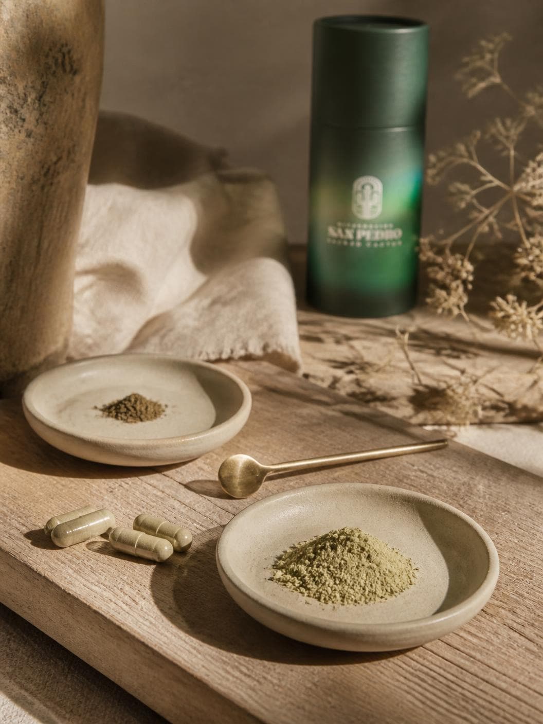 Two shallow ceramic dishes on a timber board, the nearer holding a heap of pale sage-green milled powder and the further a small pinch of a darker olive one, a long brass spoon between them, three pale capsules loose on the wood and the green tube standing out of focus behind.